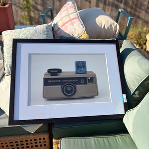 May include: A framed print featuring a tan Kodak Hawkeye Instamatic II camera with a black lens and a blue flash cube on top. The camera body displays the text HAWKEYE INSTAMATIC II in black lettering against a white mat and black frame.