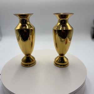 May include: A pair of matching gold-colored metal vases with a polished, reflective finish. Each vase features a slender neck, a flared rim, a rounded body, and a circular pedestal base, standing upright with a smooth, metallic surface texture.