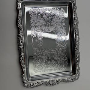 May include: A rectangular silver-toned serving tray featuring a highly reflective polished surface and an ornate, embossed border with intricate scrollwork and shell-like motifs along the edges and a decorative central panel.