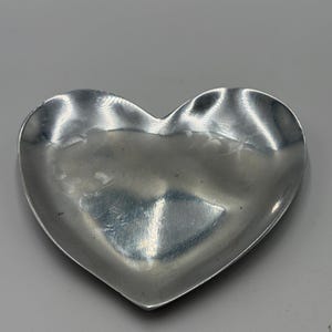 May include: A heart-shaped metal dish with a smooth, reflective silver-toned surface. The bowl features a slightly irregular, organic rim and a concave center, creating a shallow basin. The metallic finish shows subtle light reflections throughout.