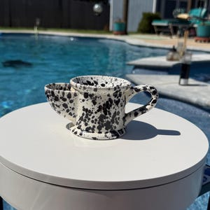 Puede incluir: Un cuenco de afeitar de cerámica con base blanca y patrón de manchas negras. El recipiente cuenta con un cuenco principal ancho para hacer espuma, un depósito lateral más pequeño para el cepillo de afeitar y un asa curva en el lateral.
