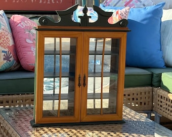 May include: A rectangular wooden display cabinet with two glass-paned doors, featuring a natural light brown wood finish on the frame and a contrasting black decorative top piece with a central finial and two small black metal handles.