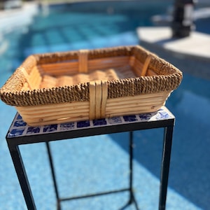 May include: A square woven basket with a light tan base and a dark brown braided rim. The basket features vertical support strips along the sides and a flat, woven bottom, providing a structured container for storage or display purposes.