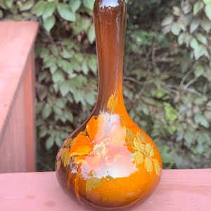 May include: A bulbous glass vase with a long, slender neck, featuring a gradient of deep amber to light orange tones. The body of the vase is decorated with a hand-painted floral design in shades of orange and yellow with green leaves.