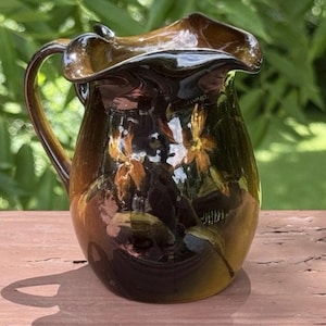 May include: A dark brown ceramic pitcher with a glossy, reflective finish and a ruffled, wavy rim. The body of the pitcher features hand-painted orange and yellow floral motifs with green leaves, complemented by a curved handle on the left side.