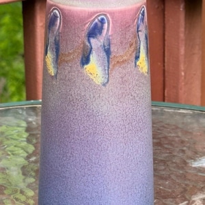 Könnte beinhalten: Eine zylindrische Keramikvase mit einer Farbverlaufglasur, die von einem tiefen Blau an der Basis zu einem sanften Violett und Rosa am oberen Rand übergeht. Der obere Bereich ist mit einer Reihe erhabener, tropfenförmiger blau-gelber Akzente verziert.