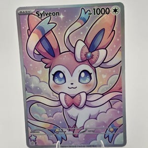 Puede incluir: Una tarjeta coleccionable rectangular que presenta al personaje Sylveon en rosa y blanco con grandes ojos azules, un lazo y antenas en forma de cinta. La tarjeta muestra el texto Sylveon, 1000 HP y basic sobre un fondo pastel de nubes y estrellas.