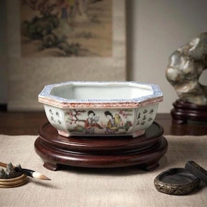 May include: An octagonal ceramic planter featuring hand-painted figures in traditional attire, surrounded by floral motifs and black Chinese calligraphy. The rim is decorated with blue and red geometric patterns, and the vessel rests on a dark wood stand.