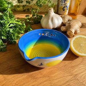 May include: A ceramic grater bowl featuring a vibrant blue and yellow gradient interior with a textured center for grating. The exterior is white with a painted yellow lemon motif and green leaves. It includes a small pouring spout and three holes.