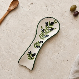May include: A ceramic spoon rest with a cream-coloured base and a dark green rim. The interior features a hand-painted design of green olive branches with black olives. The spoon rest has an elongated, curved shape designed to hold a kitchen spoon.
