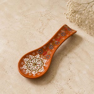 May include: A terracotta spoon rest featuring a hand-painted white mandala design in the center of the bowl, complemented by smaller blue and red floral motifs that extend along the elongated handle, all rendered in a vibrant, detailed pattern.