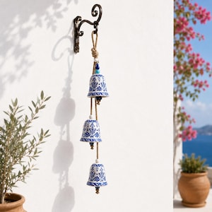 May include: A set of three ceramic bells with a white and blue floral pattern, suspended vertically from a natural jute rope. The bells are attached to a dark metal wall-mounted hook, featuring small blue and white accent beads between each bell.