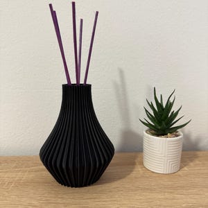 Peut inclure: Un vase noir nervuré au col effilé contenant quatre fins bâtonnets diffuseurs violets, placé à côté d'un petit pot cylindrique blanc orné d'un motif géométrique en relief et d'une plante succulente verte aux feuilles pointues.