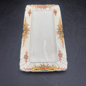 May include: A rectangular ceramic serving platter with a cream-colored center and a wide border featuring a floral pattern in shades of orange, green, and yellow. The edges are scalloped with black line accents and thin radiating black lines.
