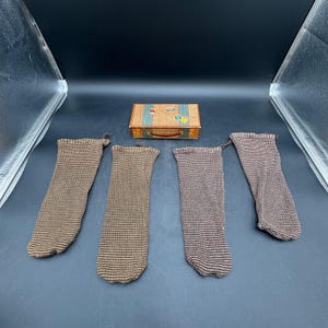 May include: Four brown and tan striped knit stockings arranged in a row, accompanied by a small rectangular box designed to resemble a vintage suitcase with a handle and colorful travel-themed stickers on the top surface.