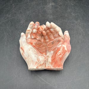 May include: A decorative bowl shaped like two cupped hands joined together. The object is crafted from a smooth, polished stone material featuring a marbled pattern with swirling veins of deep red, orange, and creamy white colors throughout.
