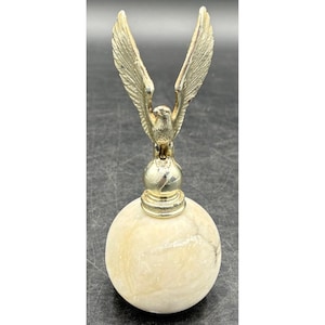 May include: A decorative object featuring a polished, cream-colored marble sphere base topped with a metallic gold-toned eagle figurine. The eagle is depicted with its wings fully extended upward, perched atop a smaller, smooth, spherical gold-toned pedestal.