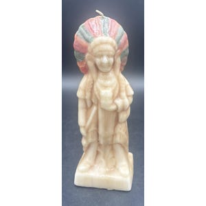 May include: A molded wax candle shaped like a figure wearing a traditional headdress with red, green, and white feathers. The figure is standing on a rectangular base, rendered in a light cream color with a single wick protruding from the top.