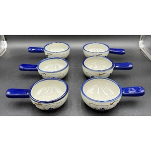 May include: A set of six small ceramic bowls with handles, each featuring a cream-colored base with hand-painted blue floral accents and a solid blue handle. The bowls are arranged in two columns of three, showcasing a consistent design.