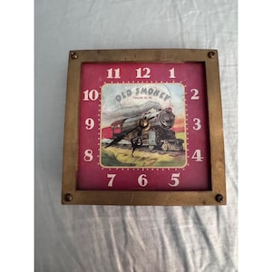 May include: A square wall clock with a metallic brass-toned frame secured by four corner screws. The clock face features a magenta background with white numerals and a central illustration of a steam locomotive labeled Old Smokey Engine No 18.