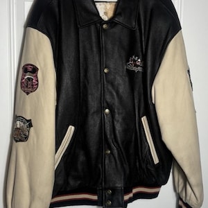 May include: A black leather varsity jacket with cream-colored sleeves, featuring a Walt Disney World logo on the chest, embroidered patches on the right sleeve, a button-front closure, and a patterned interior lining with a map design.
