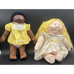 May include: Two soft-sculpture dolls with yarn hair. The left doll has dark skin, black yarn hair, and a yellow dress with white trim. The right doll has light skin, yellow yarn hair, and a white dress with a yellow pattern and a yellow ribbon tie.