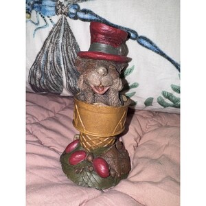 May include: A brown bear figurine wearing a red top hat with a grey band, emerging from a tan waffle cone. The base is decorated with green leaves, small brown acorns, and several smooth red berries, all crafted with a textured, painted finish.
