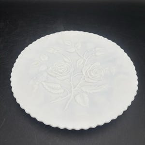 May include: A round white milk glass plate featuring a raised, embossed design of two blooming roses with leaves and stems in the center. The plate has a scalloped edge and a smooth, opaque finish, showcasing a detailed floral relief pattern.