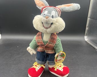 May include: A plush rabbit toy with long gray ears, wearing a green and white striped long-sleeve shirt, a brown plaid vest, blue denim shorts with rolled cuffs, and red sneakers with yellow laces. A circular tag with Bugs Bunny is attached.