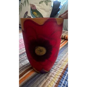 May include: A cylindrical ceramic vase featuring a hand-painted large red flower with a dark center and visible brushstrokes. The upper rim of the vase is a natural tan color, while the body is dominated by the vibrant red floral design.