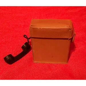 May include: A small, rectangular, tan-colored plastic toy telephone base with a hinged lid. Attached to the side of the base by a short black cord is a miniature black telephone handset. The object is constructed from smooth, solid-colored plastic.