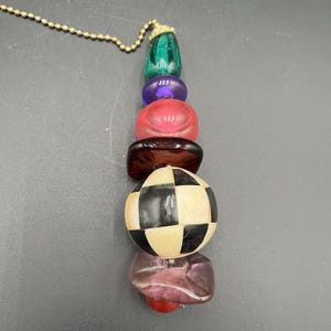 May include: A decorative pull chain featuring a vertical arrangement of multi-colored beads. The beads include a green teardrop, a purple disc, a red disc, a dark brown disc, a large black and white checkered sphere, and a translucent purple base.