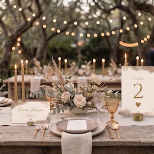 Op de afbeelding: Een bruiloftstafelsetting met een crèmekleurige tafelnummerkaart met de tekst Table 2 en een hartje, een naamkaartje met de tekst Emma Johnson Table 2, gouden dinerkaarsen, amberkleurig glaswerk en droogbloemstukken als tafeldecoratie.