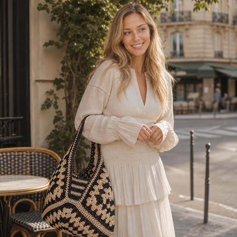 May include: A cream-coloured long-sleeved dress with a V-neckline and a tiered skirt, paired with a large black and beige crocheted hobo bag featuring a geometric pattern. The bag has a wide shoulder strap and a spacious, slouchy silhouette.