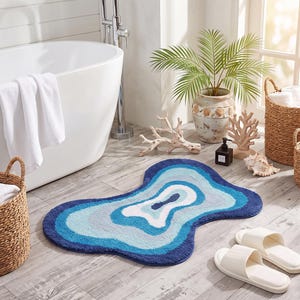 May include: An irregular, organic-shaped bath mat featuring concentric layers of blue, light blue, and white colors. The rug has a textured, tufted surface and a wavy, fluid outline that mimics the appearance of a cross-section of a stone.