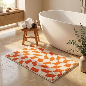 Può includere: Un tappetino da bagno rettangolare caratterizzato da un vivace motivo a scacchi arancione e bianco con un design a griglia ondulata e distorta. Il tappeto ha una consistenza spessa e trapuntata e poggia su un pavimento piastrellato chiaro.