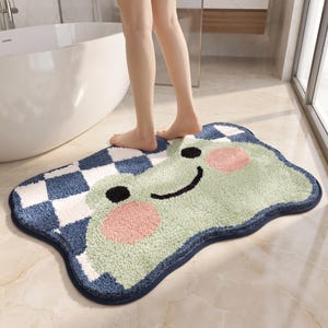 May include: A rectangular bath mat with rounded edges featuring a light green frog face with two pink circular cheeks, black eyes, and a black smile. The remaining area displays a blue and white checkered pattern with a dark blue border trim.