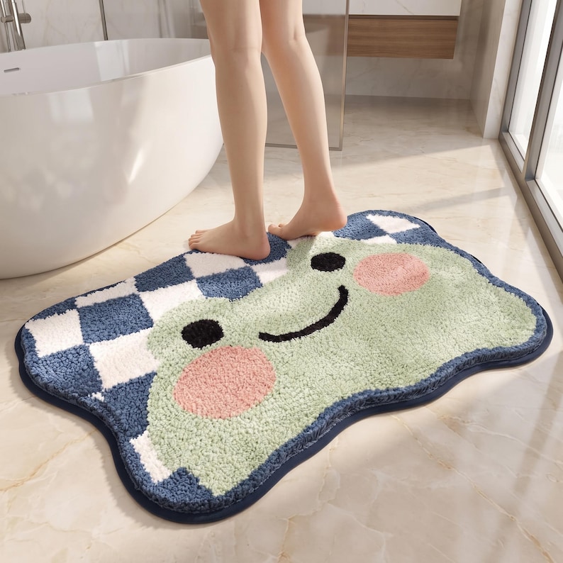 May include: A rectangular bath mat with rounded edges featuring a light green frog face with two pink circular cheeks, black eyes, and a black smile. The remaining area displays a blue and white checkered pattern with a dark blue border trim.