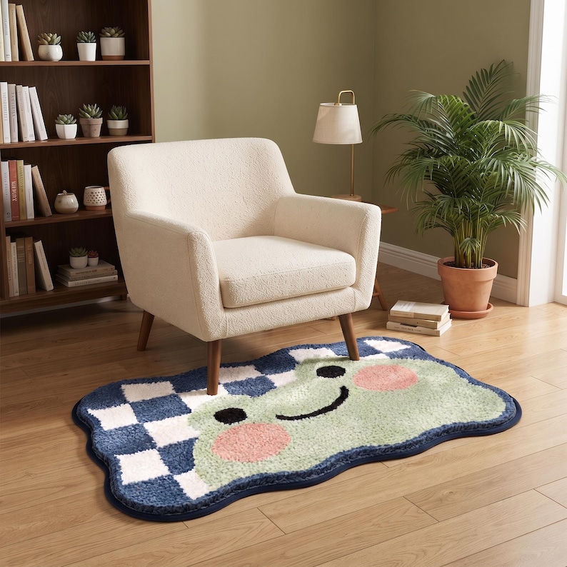 May include: A rectangular rug with rounded, wavy edges featuring a light green frog face with pink cheeks and black eyes. The background pattern consists of a blue and white checkerboard design. The rug has a dark blue border trim throughout.