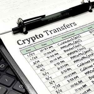 May include: A black ballpoint pen rests on a printed ledger titled Crypto Transfers, which features columns for Date, Time, From Account, Asset, and Amount. The document is held in a black binder next to a black computer keyboard with visible keys.