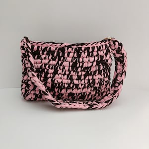May include: A rectangular crochet handbag crafted from thick, intertwined fabric yarn in alternating horizontal stripes of black and light pink. The bag features a braided shoulder strap made from the same black and pink yarn, attached to the top corners.