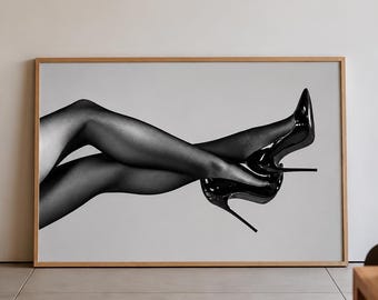 May include: A black and white framed wall art print featuring a pair of legs wearing sheer black pantyhose and glossy black high-heeled stiletto pumps. The legs are positioned horizontally with the knees bent and feet crossed in the air.