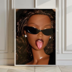 May include: A framed wall art print featuring a close-up portrait of a doll with dark brown skin, long wavy dark brown hair, and black cat-eye sunglasses. The doll has gold eyeshadow, dark lipstick, and a pink tongue sticking out, wearing small stud earrings.