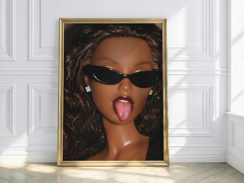 May include: A framed wall art print featuring a doll with dark brown skin, long wavy brown hair, and dark red lipstick. The doll wears black oval sunglasses and small square stud earrings, with its tongue sticking out in a playful expression.