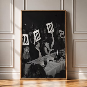 Peut inclure: Une photographie encadrée en noir et blanc montrant un groupe de personnes attablées, dont plusieurs tiennent des panneaux blancs affichant le chiffre 10. La scène comprend des verres, une bouteille et une nappe blanche dans une ambiance tamisée.