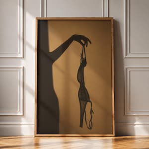 Puede incluir: Una impresión de arte mural enmarcada que presenta la silueta oscura de una mano sosteniendo una prenda de lencería sobre un fondo de color tostado sólido. La obra muestra la sombra de un brazo y una mano que cuelga una prenda delicada con tirantes finos.