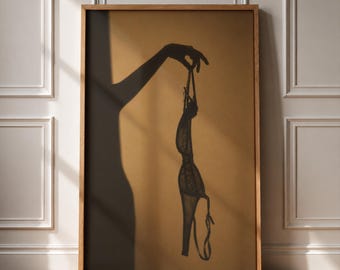 May include: A framed wall art print featuring a dark silhouette of a hand holding a piece of lingerie against a solid tan background. The artwork displays the shadow of an arm and a hand dangling a delicate garment with thin straps and lace details.