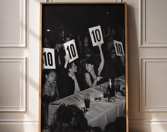 May include: A framed black and white photograph depicting a group of people seated at a table, with several individuals holding up white signs displaying the number 10. The scene includes glassware, a bottle, and a white tablecloth in a dimly lit setting.