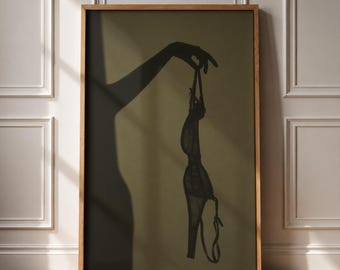 May include: A framed wall art print featuring a dark silhouette of an arm and hand holding up a lacy brassiere against a muted olive green background. The artwork displays a high-contrast shadow effect, emphasizing the delicate texture of the lingerie.
