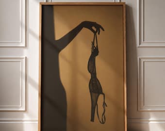 May include: A framed wall art print featuring a dark silhouette of an arm and hand holding a piece of black lace lingerie against a solid tan background. The artwork displays a high-contrast composition with clean lines and a minimalist aesthetic.